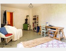 Ein schlicht eingerichtetes Zimmer mit hellem Anstrich und Holzboden. Der Raum enthält einen Schreibtisch mit Nähmaschine, ein Bücherregal, einen grünen Ofen, eine Sitzecke mit Polstermöbeln und eine offene Tür, die zu einem angrenzenden Raum mit warmen Wandfarben führt.