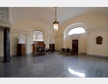 Geräumiger Innenraum mit einer hohen, gewölbten Decke und einem glänzenden Marmorboden. Der Raum verfügt über dekorative Elemente wie Säulen aus schwarzem Marmor, weiße Statuen auf Holzsockeln, einen Holz-Kamin und große Bogenfenster, die natürliches Licht hereinlassen; mehrere Tische und Stühle sind symmetrisch angeordnet.