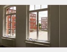Blick aus großen Fenstern eines Gebäudes im Innenraum auf einen gepflasterten Hof mit roten Backsteingebäuden und modernen Ziegelbauten im Hintergrund. Unter den Fenstern befinden sich Heizkörper.