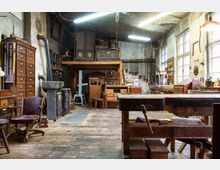 Eine traditionelle Schreinerwerkstatt mit Holzböden, großen Werkbänken, vielen Werkzeugen an den Wänden und Schränken aus Holz. Der Raum ist mit natürlichen Licht durch große Fenster beleuchtet, während Neonleuchten an der Decke hängen.