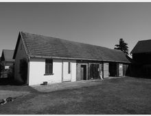Schwarz-Weiß-Fotografie eines einstöckigen ländlichen Gebäudes mit Satteldach und einer Fassade aus verputztem Mauerwerk sowie Holztüren und -fenstern. Im Vordergrund liegt eine grasbewachsene Fläche, und rechts sind weitere landwirtschaftliche Nebengebäude erkennbar.