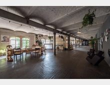 Weitläufiger Innenraum eines Industrielofts mit sichtbaren Backsteindecken, großen Fensterbögen und vereinzelten Stahlträgern. Der Raum ist mit Holztischen, unterschiedlich gestalteten Stühlen, hängenden Pflanzen und dekorativen Platten an den Wänden ausgestattet; eine Bühne mit Musikinstrumenten ist ebenfalls sichtbar.