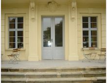 Gelbe Fassade eines historischen Gebäudes mit einer verzierten Doppeltür aus Glas in der Mitte, flankiert von zwei Fenstern mit weißen Sprossen. Vor der Tür stehen mehrere Klappstühle und -tische auf einer gepflasterten Terrasse, die über eine kleine Steintreppe erreichbar ist.