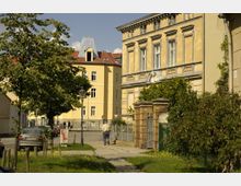 Straßenszene in einem Wohngebiet mit historischen Gebäuden, darunter ein gelb gestrichenes Haus mit Giebel und ein verziertes Gebäude mit Ornamenten. Im Vordergrund ein Gehweg, umgeben von Bäumen, parkenden Autos und einem Ziegelzaun mit einem Metalltor.