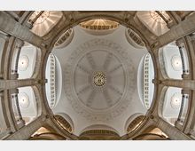 Blick von unten auf eine prächtige Kuppel im Innenraum eines historischen Gebäudes, verziert mit detaillierten Stuckelementen und einem zentralen Kronleuchter. Rundbögen, Balustraden und helle Wände prägen die Architektur.