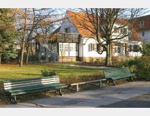 Grüne Parkbänke stehen auf einem gepflasterten Weg vor einem weißen Gebäude mit rotem Satteldach und großen Fenstern, umgeben von Bäumen und einem Rasenbereich. Eine Außentreppe führt zu einem Balkon an einer Seite des Hauses.