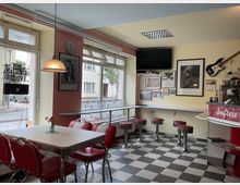 Innenansicht eines kleinen Diners mit schwarz-weiß kariertem Boden, roten Sitzbänken und Chrom-Barhockern an einer Fensterbar. An der Wand hängen gerahmte Bilder und eine E-Gitarre, daneben steht eine rote Theke mit der Aufschrift „Softeis“ und großen Fenstern zur Straße.