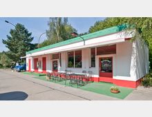 Ein kleines Café mit rechteckigem, einstöckigem Gebäude, weißer Fassade und rotem Sockel, einem begrünten Flachdach und großen Fenstern. Vor dem Eingang stehen schwarze Metalltische und -stühle auf einer gepflasterten Terrasse, umgeben von Bäumen und einem parkenden blauen Lieferwagen.