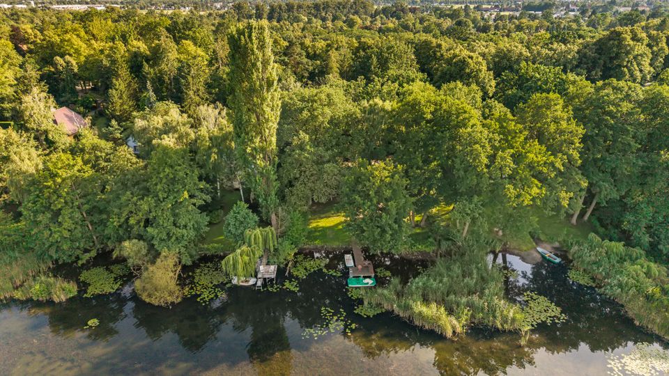 Luftaufnahme eines bewaldeten See- oder Flussufers mit dichtem Baumbestand, Schilf- und Seerosenflächen im flachen Wasser. Am Ufer liegen mehrere kleine Stege mit Booten, im Hintergrund erstreckt sich eine flache Landschaft mit vereinzelter Bebauung.