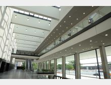 Großes, modernes Atrium mit mehreren Etagen, einem Glasgeländer entlang der oberen Etage und großen Fenstern, die viel Tageslicht hereinlassen. In der Mitte führt eine Rolltreppe nach oben, während die Decke von hängenden Lampen beleuchtet wird.