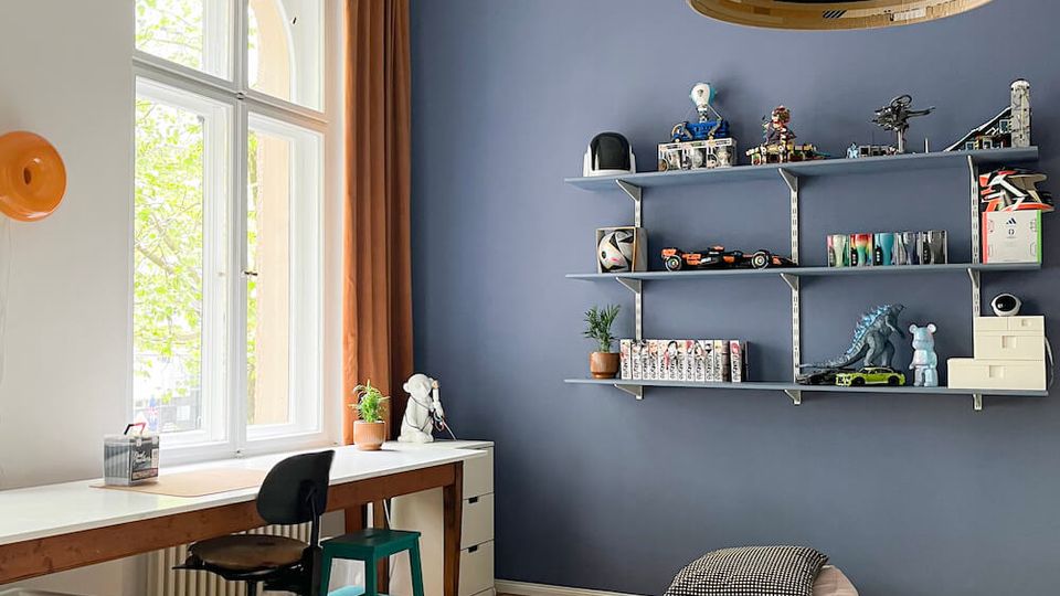 Bright study room with a tall arched window, ornate white ceiling molding, and a blue accent wall with long shelves displaying toys and books. A long desk with chairs sits by the window, and a light rug with a small low table and floor cushion covers the wooden floor.