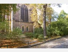 Außenansicht eines historischen Backsteingebäudes mit großen, spitzbogigen Fenstern, umgeben von Laubbäumen und herbstlichem Blattwerk; im Vordergrund ein gepflasterter Gehweg und Stufen.