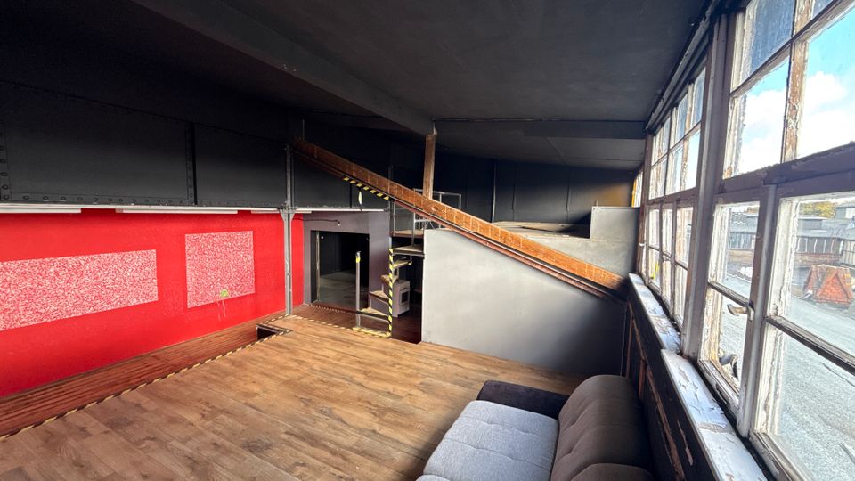 Industrieartiger Innenraum mit Holzfußboden, roter Wandfläche und schwarzer Stahlkonstruktion sowie einer schrägen Rampe zu einer erhöhten Ebene. Rechts verläuft eine Reihe großer, alter Sprossenfenster mit Blick auf die umliegenden Dächer.