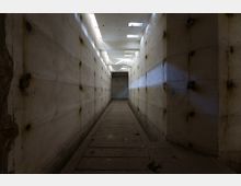 Ein schmaler, leerer Betonflur mit hohen Wänden zeigt sichtbare Verschraubungen und Belagsrückstände. Oberlichter an der Decke lassen Tageslicht in den Raum, wodurch diagonale Lichtstrahlen entstehen.