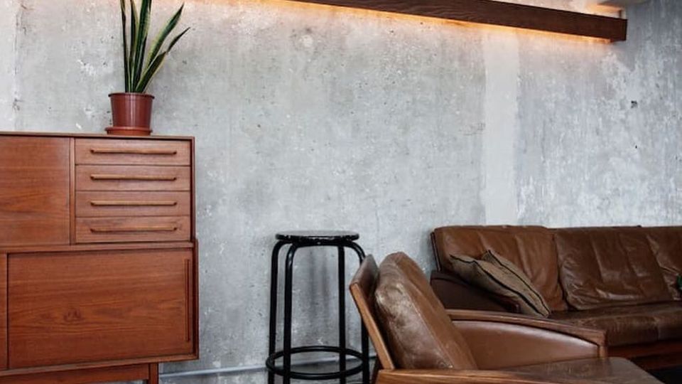 Industrial anmutender Wohnraum mit rauer Betonwand und einer langen Holzleiste mit indirekter Beleuchtung. Im Vordergrund stehen ein braunes Ledersofa und ein Ledersessel, daneben ein schwarzer Barhocker und ein Holzsideboard mit Topfpflanze.