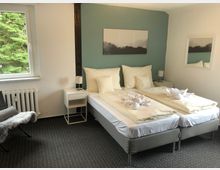 Helles Schlafzimmer mit zwei Einzelbetten, die zu einem Doppelbett zusammengestellt sind, dekoriert mit gefalteten Handtüchern. Links ein Fenster mit Blick auf Bäume, rechts ein Nachttisch mit Lampe; der Teppichboden ist grau, die Wand hinter dem Bett in Grün-Blau gestrichen.