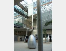 Modernes Innenraumdesign eines Büro- oder Bildungsgebäudes mit Sichtbetonwänden, einem Glasdach und mehreren offenen Etagen, die durch Treppen und Brücken verbunden sind. Im Vordergrund stehen futuristisch geformte, silberne Arbeitskabinen, während eine Palme und Glasflächen eine helle und offene Atmosphäre schaffen.