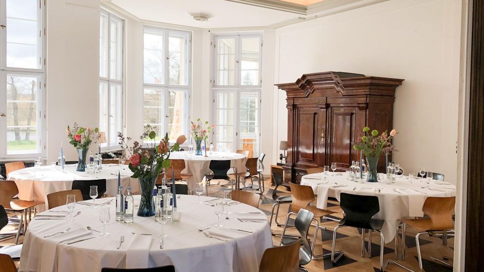 Heller Bankettsaal mit großen Sprossenfenstern und Stuckdecke, ausgestattet mit runden Tischen mit weißen Tischdecken, Gedecken und Blumenvasen. An der Wand steht ein großer dunkler Holzschrank; der Boden ist ein Parkett im geometrischen Muster.