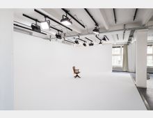 Ein minimalistisches Fotostudio mit weißem, nahtlosem Hintergrund und Deckenbeleuchtung aus mehreren Scheinwerfern auf Schienen. In der Mitte steht ein einzelner Holzstuhl, und im Hintergrund sind große Fenster mit Vorhängen zu sehen.