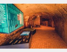 Ausstellungsraum in einem historischen Gewölbekeller mit Backsteinmauern und -boden. Links zeigt eine Tafel mit Texten und Bildern eine Ausstellung, während im Hintergrund gestapelte Ziegelsteine und eine Schubkarre sichtbar sind.