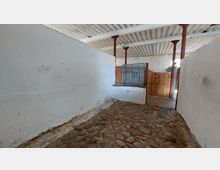 Innenansicht eines einfachen Stalls mit weiß gestrichenen Wänden, einer gepflasterten Bodenfläche und einer Holzbox mit Metallgitter. Rote Metallstützpfeiler und eine Holztür sind im Hintergrund zu sehen.