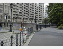 Eine städtische Szene mit gepflastertem Gehweg, metallenen Pollern und laternenartigen Straßenlampen; im Hintergrund steht ein großes, mehrstöckiges Wohngebäude mit Balkonen. Auf der rechten Seite befindet sich ein Gehweg, der von Bäumen gesäumt ist.