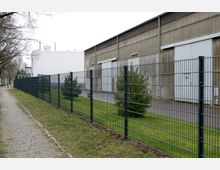 Industriegebiet mit einer großen Lagerhalle aus Wellblech und einem vorgelagerten Metallzaun. Der Bereich umfasst eine gepflasterte Gehwegseite, Grünflächen mit kleinen Nadelbäumen und asphaltierte Zufahrtswege.