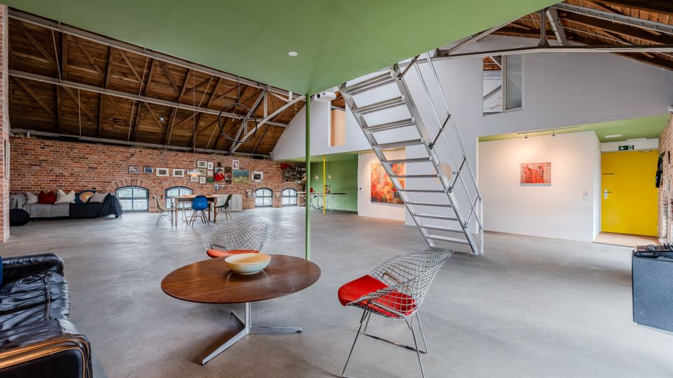 Großzügiger Loftraum in einem umgebauten Industriegebäude mit sichtbarer Holzbalkendecke, Backsteinwänden und Betonboden. Eine moderne Metalltreppe führt zu einer Galerie; im offenen Wohnbereich stehen Sitzmöbel und Tische, an den Wänden hängen Kunstwerke und es gibt mehrere niedrige Rundbogenfenster.