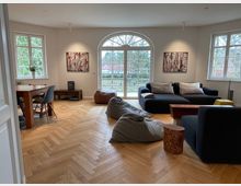 Helles Wohnzimmer mit Parkettboden und großen Fenstern, darunter eine doppelflügelige Glastür mit Blick auf einen Garten. Der Raum ist möbliert mit einer schwarzen Sofalandschaft, einem Holztisch mit Stühlen, Sitzsäcken und dekorativen Holzelementen, sowie abstrakten Gemälden an den Wänden.