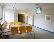 Ein Wartebereich in einem öffentlichen Gebäude mit gelben Stuhlreihen in der Mitte, weißen Wänden und mehreren Bildern an den Wänden. An der Decke hängt ein Monitor mit einer Anzeige, und auf der rechten Seite führt ein langer Flur weiter ins Gebäude.