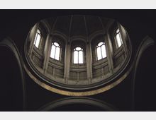 Innenansicht einer Kuppel in einer historischen Kirche mit hohen Rundbogenfenstern, schmalen Säulen und umlaufender Galerie. Der Raum ist dunkel, wobei das Licht durch die Fenster einfällt und die Architektur betont.