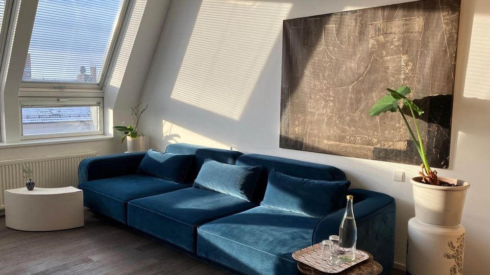 Modern living room with a large blue sectional sofa beneath a slanted ceiling and tall window with blinds casting light patterns on white walls. An abstract wall artwork, small round tables, and potted plants add decor on a dark wood floor.