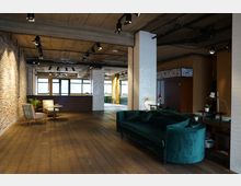 Industrieller Innenraum mit offenen Backsteinwänden, freiliegender Betondecke und Holzboden. Der Raum ist mit gemütlichen Sitzmöbeln, einer grünen Samtcouch und einem kleinen Tisch eingerichtet, während große Fenster natürliches Licht hereinlassen.