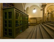 Innenansicht eines historischen Gebäudes mit einer grünen, verglasten Kabine. Der Raum ist geprägt von hohen, weißen Gewölbedecken, Steinsäulen, einer Treppe mit Geländer aus Glas sowie einem Heizkörper an der Wand.