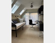 Wohnzimmer oder Arbeitsbereich in einer Dachgeschosswohnung mit schrägen Decken und Dachfenstern. Im Raum befinden sich ein Tagesbett, ein schwarzer Bürostuhl, ein kleiner Tisch, ein Regal mit Metallrahmen und ein Vorhang vor einer großen Fensterfront.