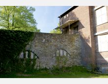 Eine Außenansicht eines Gebäudeteils mit einer Steinmauer, die zwei rundbogige Fenster mit Glasbausteinen beinhaltet und teilweise mit Efeu bewachsen ist. Daneben ist ein Backsteingebäude mit mehreren geschlossenen Fenstern und einem kleinen Balkon zu sehen, umgeben von einer grünen, bewachsenen Wiese und Bäumen im Hintergrund.