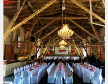 Großer Veranstaltungsraum in einem historischen Gebäude mit offenem Holzbalkendach und Backsteinwänden, dekoriert mit Lichterketten und Kronleuchtern. Die Tische sind in Reihen mit weißen Tischdecken und Stuhlhussen aufgestellt und für eine Feier eingedeckt.