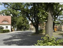 Straßenecke in einer ländlichen Umgebung mit einem kleinen Gebäude mit rotem Ziegeldach links und einer Baumreihe entlang der Straße. Im Hintergrund sind Steinmauern und weitere Häuser erkennbar, umgeben von Bäumen und Vegetation.