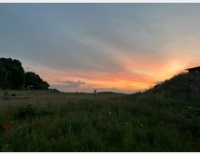 Weite Wiesenlandschaft bei Sonnenuntergang mit orangefarbenem Himmel, Mohnblumen im Vordergrund und Bäumen sowie einer kleinen Hütte am linken Bildrand. Kleidungshügel und Gebäudeecke rechts im Bild.