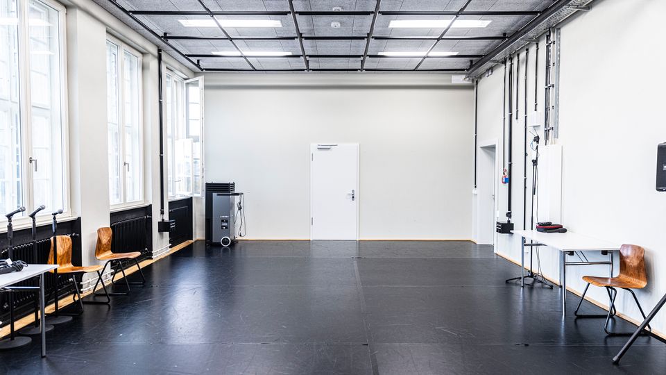 Heller, leerer Proben- oder Trainingsraum mit schwarzem Boden und weiß gestrichenen Wänden. Links befinden sich große Fenster und ein paar Stühle, rechts Kabelkanäle sowie ein Tisch; an der Stirnwand steht eine weiße Tür unter einer Rasterdecke mit rechteckigen Leuchten.