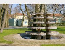 Springbrunnen aus gestapelten, ringförmigen Betonelementen in einem Park mit Rasenfläche und umgebenden Bäumen; im Hintergrund ein Gebäude mit hellgelber Fassade und rotem Dach sowie ein kleiner Pavillon.