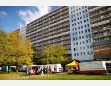 Ein mehrstöckiges Wohngebäude mit Balkonen im Hintergrund, davor ein kleiner Markt mit Verkaufsständen, darunter ein gelber Pavillon und ein Feinkostwagen. Laubbäume mit gelb gefärbten Blättern und eine Grünfläche prägen den Vordergrund.