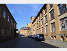 Straßenszene mit alten Industriegebäuden aus gelb-braunem Backstein, großen vergitterten Fenstern und Kopfsteinpflaster. Geparkte Autos stehen entlang der Straße, die von Graffiti-bedeckten Fassaden gesäumt ist.