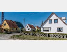 Straßenszene in einem Wohngebiet mit Einfamilienhäusern, die weiße oder gelbe Fassaden und rote oder dunkelgrüne Schrägdächer haben, umgeben von niedrigen Hecken und Zäunen aus Beton. Der Himmel ist blau, und Stromleitungen verlaufen oberirdisch entlang der Straße.