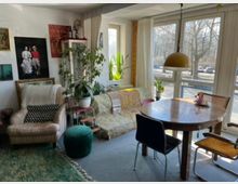 Bright living and dining area with large corner windows and sheer curtains, overlooking trees and a street outside. The room has a round wooden dining table under a dome pendant light, a patterned sofa and armchair, indoor plants, and framed artwork on white walls.