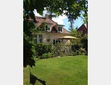 Zweistöckiges Einfamilienhaus mit hellbeiger Fassade und rotem Satteldach, umgeben von einem grünen Garten. Im Vordergrund ist eine gepflegte Rasenfläche, eine mit Steinen eingefasste Terrasse und verschiedene Sträucher sowie ein Sonnenschirm sichtbar.