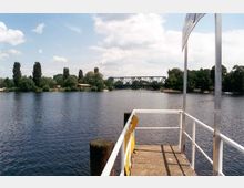 Blick von einem Steg über einen Fluss mit dicht bewachsenen Ufern, im Hintergrund eine grüne Stahlbrücke und vereinzelte Bäume unter einem teils bewölkten Himmel.
