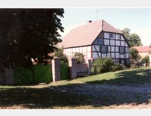 Fachwerkhaus mit rotem Ziegeldach, umgeben von einem gepflasterten Hof und einer Mauer mit grünen Toren. Im Vordergrund Rasenfläche und Bäume, im Hintergrund weitere Gebäude und blauer Himmel.