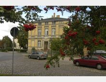 Gelbes historisches Gebäude mit verzierter Fassade, flankiert von Bäumen und roten Vogelbeeren im Vordergrund; gepflasterte Straße mit parkenden Autos im Vordergrund.