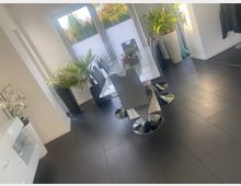 Heller moderner Essbereich mit dunkelgrauen Fliesen, einem weißen Esstisch und mehreren grauen Stühlen mit Chromfuß. Große Glastüren führen auf eine Terrasse; mehrere große Zimmerpflanzen in hohen Töpfen stehen entlang der Fensterfront.